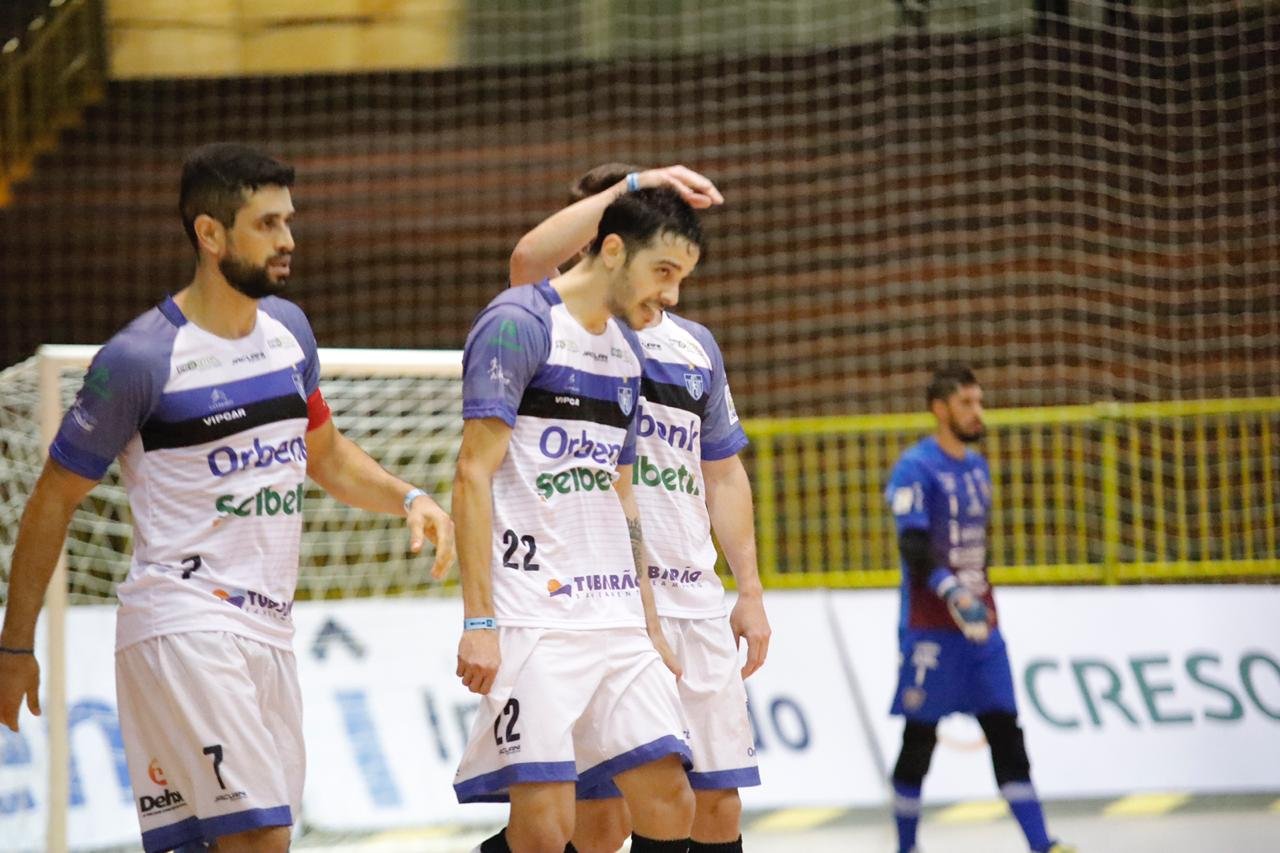 Tubarão Futsal #Pracegover Foto: Na imagem há três atletas do Tubarão Futsal e ao fundo um jogador do Campo Mourão em uma quadra de esportes