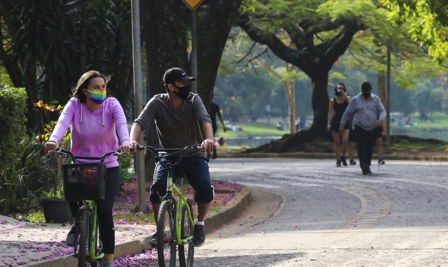 covid_ibirapuera #Pracegover Foto: na imagem há uma rua asfaltada, árvores, pessoas caminhando e duas pessoas andando de bicicleta