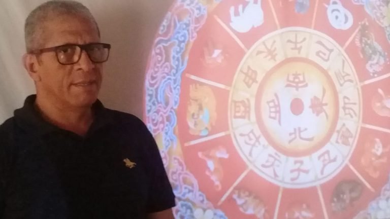 #Pracegover Foto: na imagem há um homem de camisa preta e óculos e próximo a ele um painel com os astros