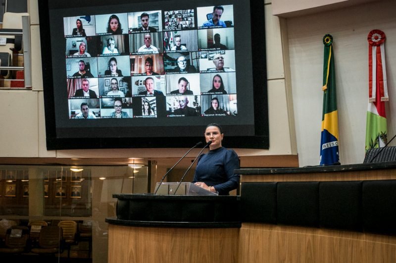 Agência AL (1) #Pracegover Foto: na imagem há uma mulher falando ao microfone e em um telão há muitas pessoas participando do ato