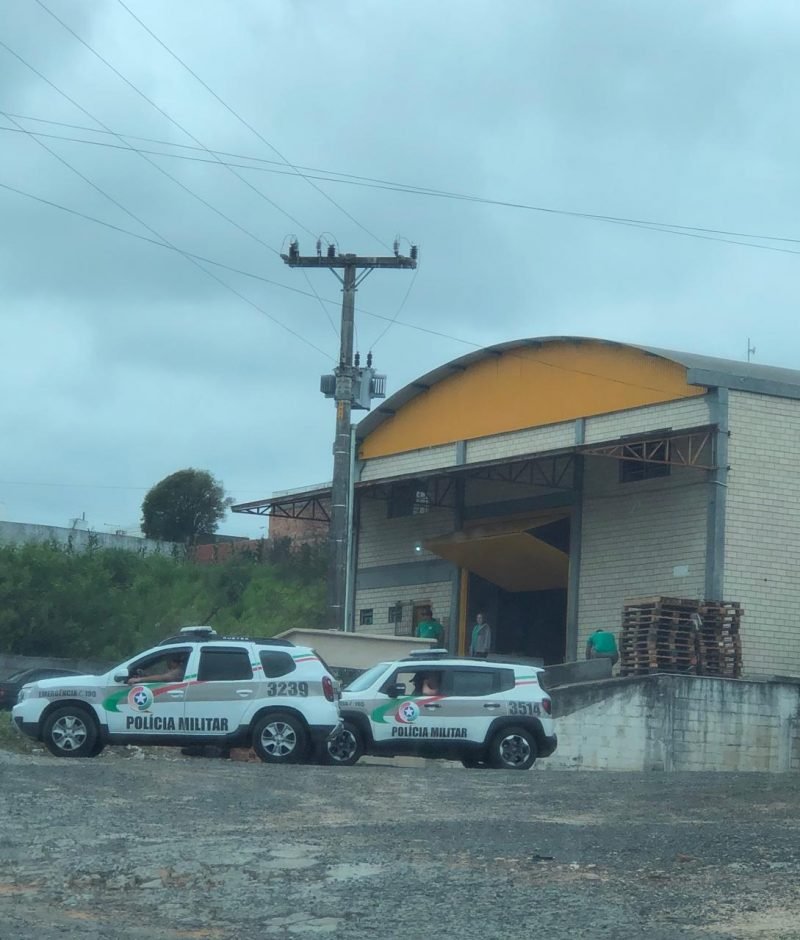 galpão #Pracegover Foto: na imagem há um galpão, duas pessoas próximas a entrada do local, duas viaturas, um carro preto e estrados