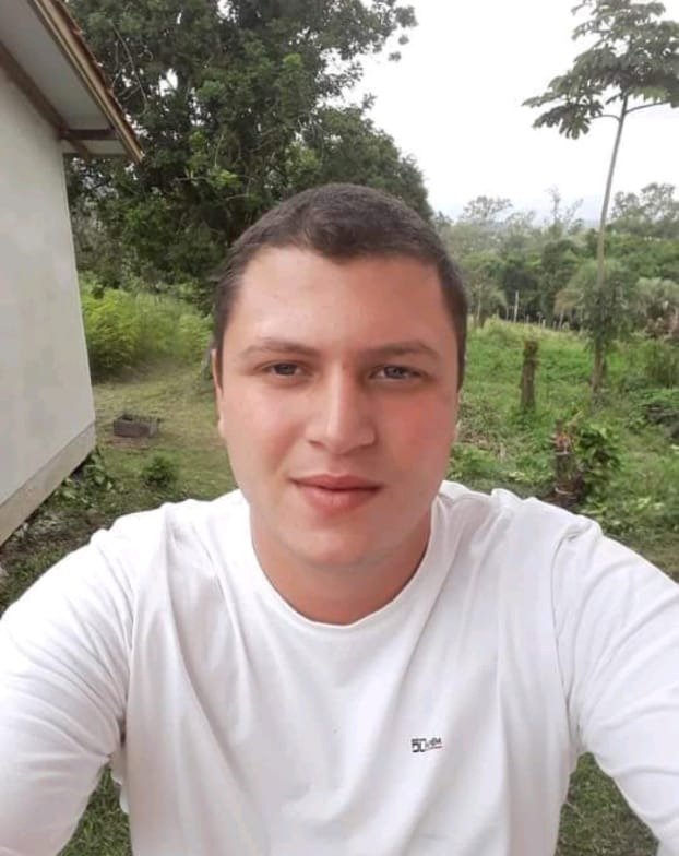 juan (1) #Pracegover Foto: na imagem há um jovem de camiseta branca. Ele está em um terreno com área verde e ao seu lado uma casa