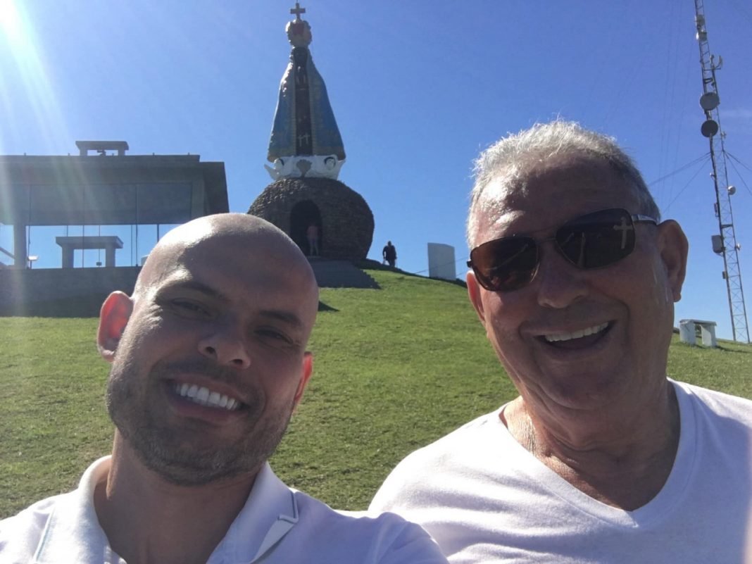 Julio #Pracegover Foto: na imagem há dois homens sorridentes e no fundo há também há imagem de Nossa Senhora Aparecida