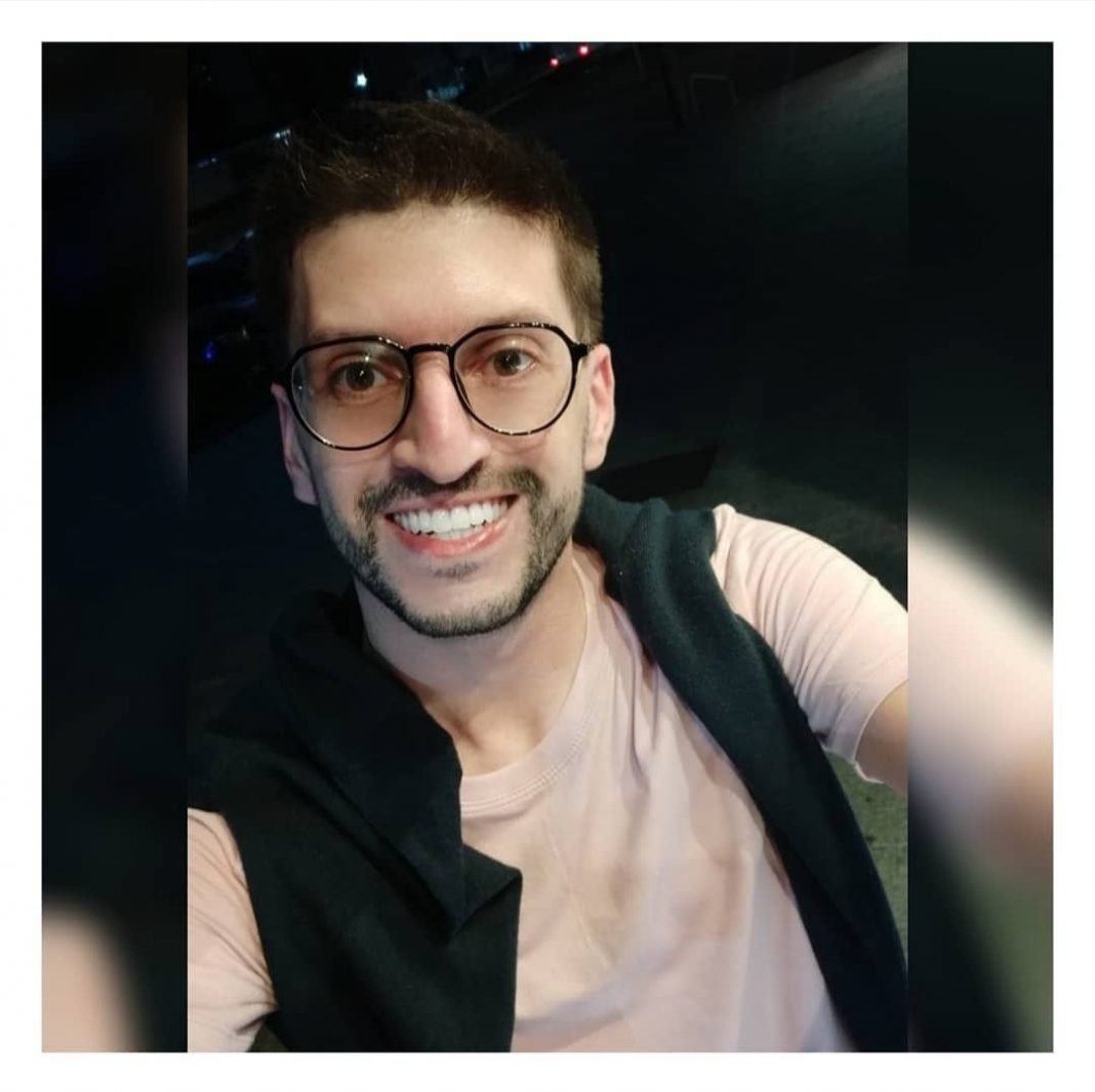 Ademir Junior #Pracegover Foto: na imagem há um jovem sorridente, de óculos de grau, camiseta clara e também com um agasalho escuro nos ombros