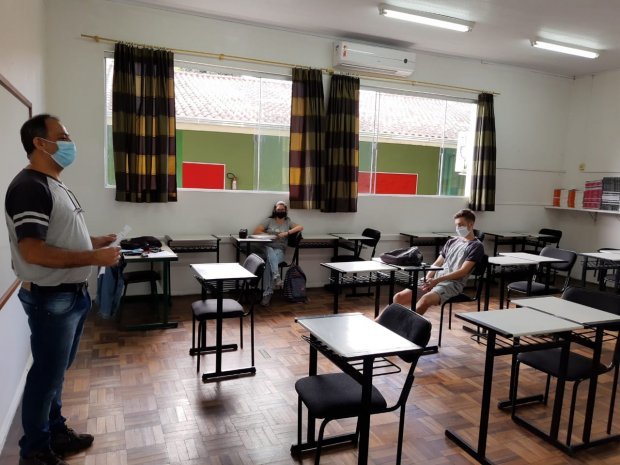 comeca_apoio_pedagogico_presencial-1 #Pracegover Foto: na imagem há uma sala de aula com várias carteiras, cortinas, janelas, um professor e dois alunos