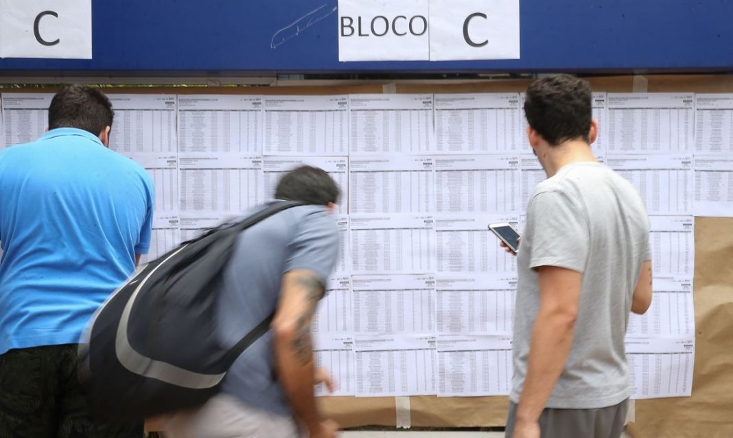 enem #Pracegover Foto: na imagem há três pessoas, conferindo uma lista. Uma delas está com um celular na mão e a outra com mochila nas costas