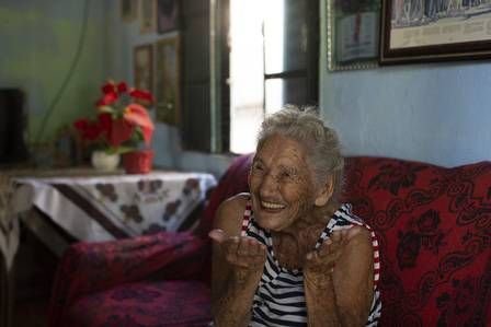 Hilda #Pracegover Foto: na imagem há uma idosa, sofa, uma mesa e janela