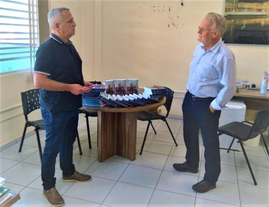 anita #Pracegover Foto: na imagem há dois homens, uma mesa, cadeirasum quadro e livros na mesa