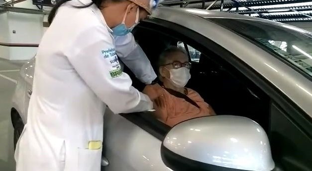 Carmen Mendes Stapazzoli, de 92 anos, é a primeira idosa a se imunizar na 2ª etapa de vacinação contra Covid-19 #pracegover Na foto, Carmem está sentada no banco do carro e uma enfermeira aplica a vacina