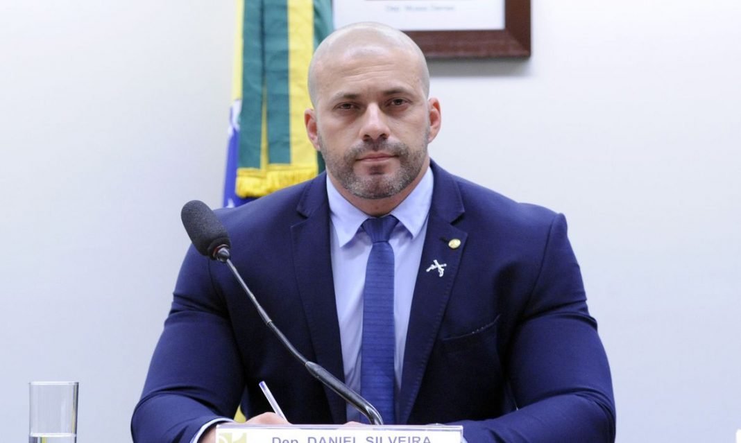 Daniel #Pracegover Foto: na imagem há um homem de terno azul, camisa branca e gravata azul. Há um microfone, uma bandeira do Brasil e um quadro