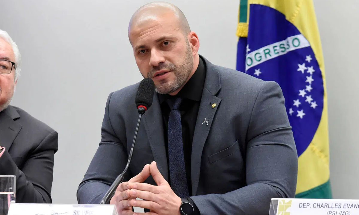 DS #Pracegover Foto: na imagem há um homem de terno escuro, sentado e uma bandeira do Brasil
