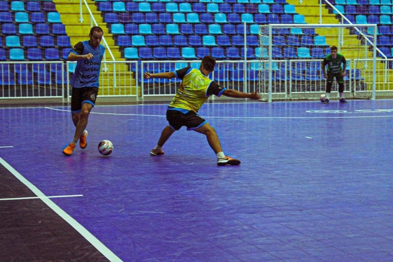 futsal #Pracegover Foto: na imagem há três atletas, uma bola, uma trave e arquibancada
