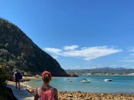 #pracegover uma mulher caminha sobre um deck entre um morro e as pedras na beira do mar