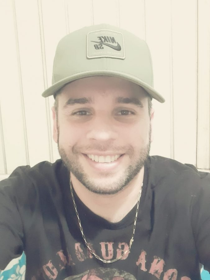 Moisés #Pracegover Foto: na imagem há um homem sorridente, de barba, com uma camiseta preta, corrente no pescoço e um boné claro