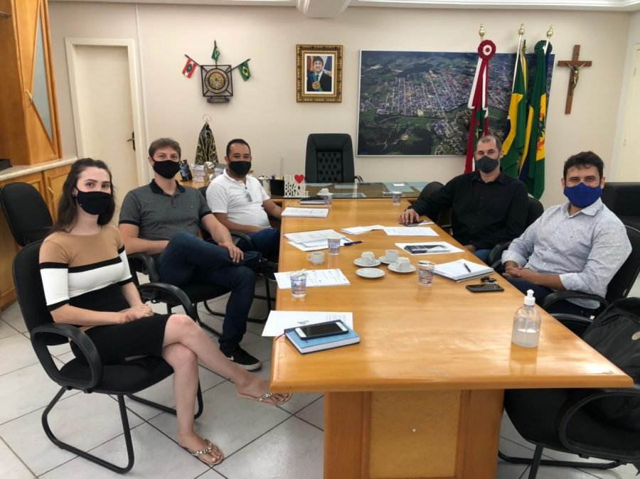 Reurb #Pracegover Foto:na imagem há pessoas sentadas, uma mesa, cadeiras, folhas e um frasco de álcool em gel