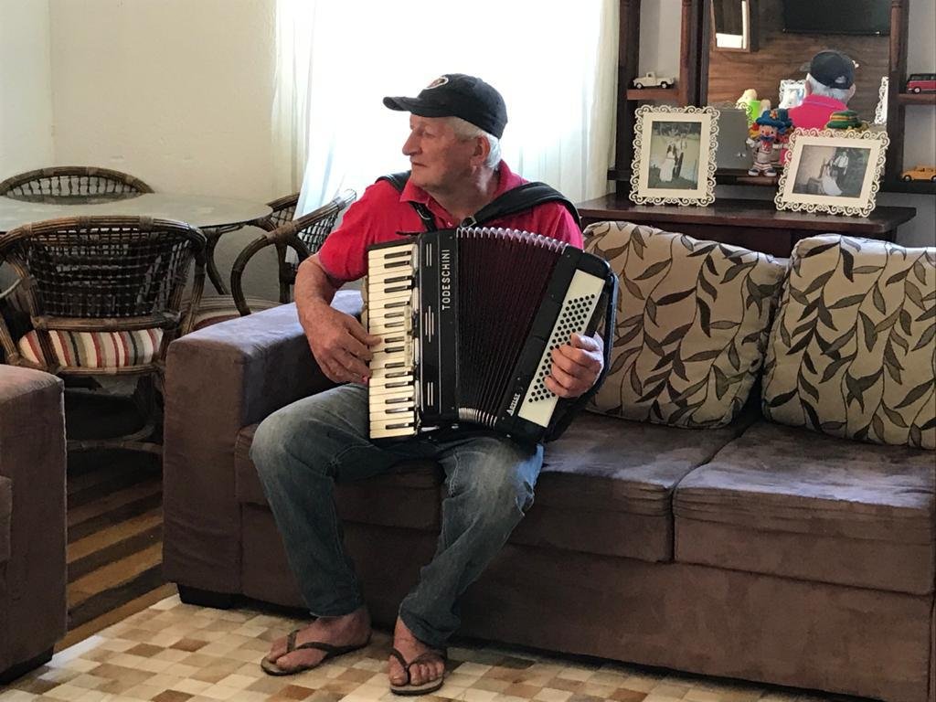 Ademar #Pracegover Foto: na imagem há um homem de boné, uma gaita, sofá, almofadas e porta retratos