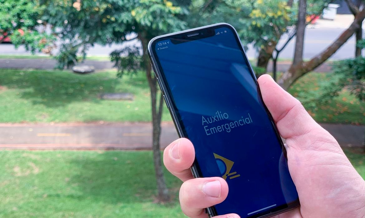 auxílio #Pracegover Foto: na imagem há uma mão, um celular, gramado e árvores