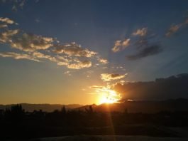 #Pracegover Na foto, céu com poucas nuvens e sol se pondo atrás de um morro