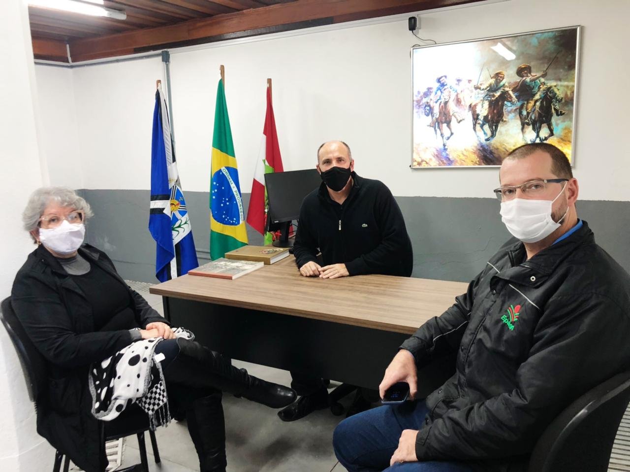 cultura tb #Pacegover Foto: na imagem há três pessoas, dois homens e uma mulher , três bandeiras, três cadeiras e uma mesa