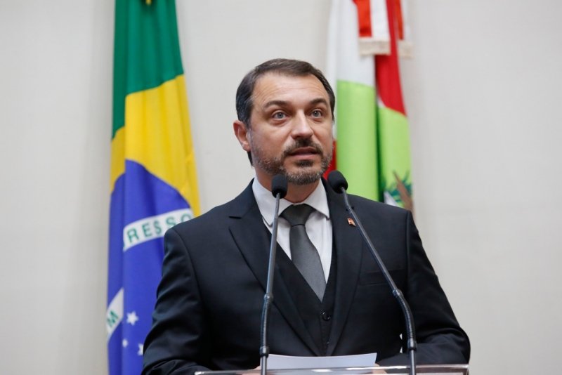 moises-1 #Pracegover Foto: na imagem há um homem de terno, um microfone e duas bandeiras, a do país e do Estado de SC