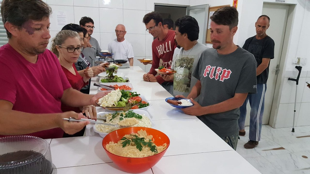O Albergue Pousada da Paz conta com quase 150 voluntarios que se revezam todas as noites para preparar a melhor janta pra todos... #Pracegover Na foto, pessoas em volta de uma mesa cheia de comida