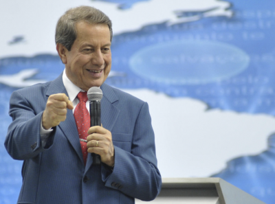 pastor #Pracegover Foto: na imagem há um homem de terno azul, camisa branca e gravata vermelha. Em uma das mãos ele segura um microfone