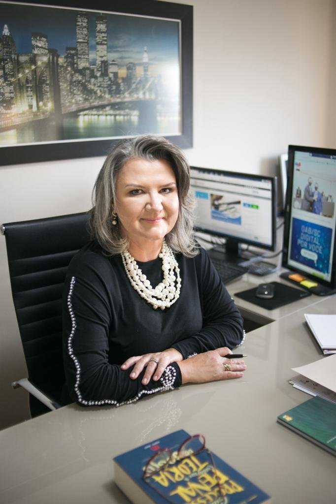 patricia #Pracegover Foto: na imagem há uma mulher, uma cadeira, mesa, computador, folhas e um livro