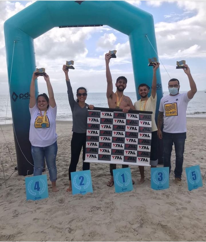 natação atn #Pracegover foto: na imagem há cinco pessoas, placas de números, o mar e areia