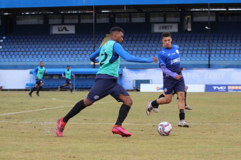 Treino #Pracegover foto: na imagem há dois atletas, um campo, uma bola e arquibancada