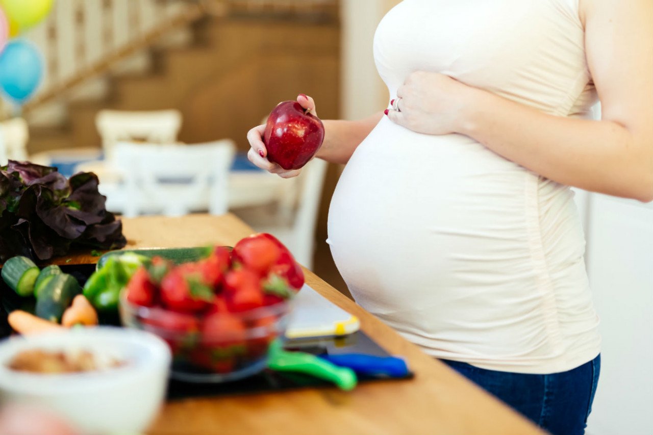 alimentação #Pracegover foto? na imagem há uma mulher grávida com a mão na barriga, na outra mão uma maça e frutas na mesa
