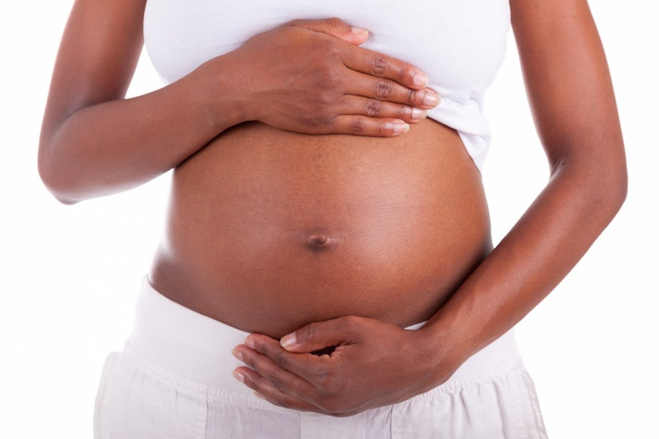 gestante #Pracegover foto: na imagem há uma mulher com as duas mães na barriga. Ela está gestante. Suas roupas são brancas