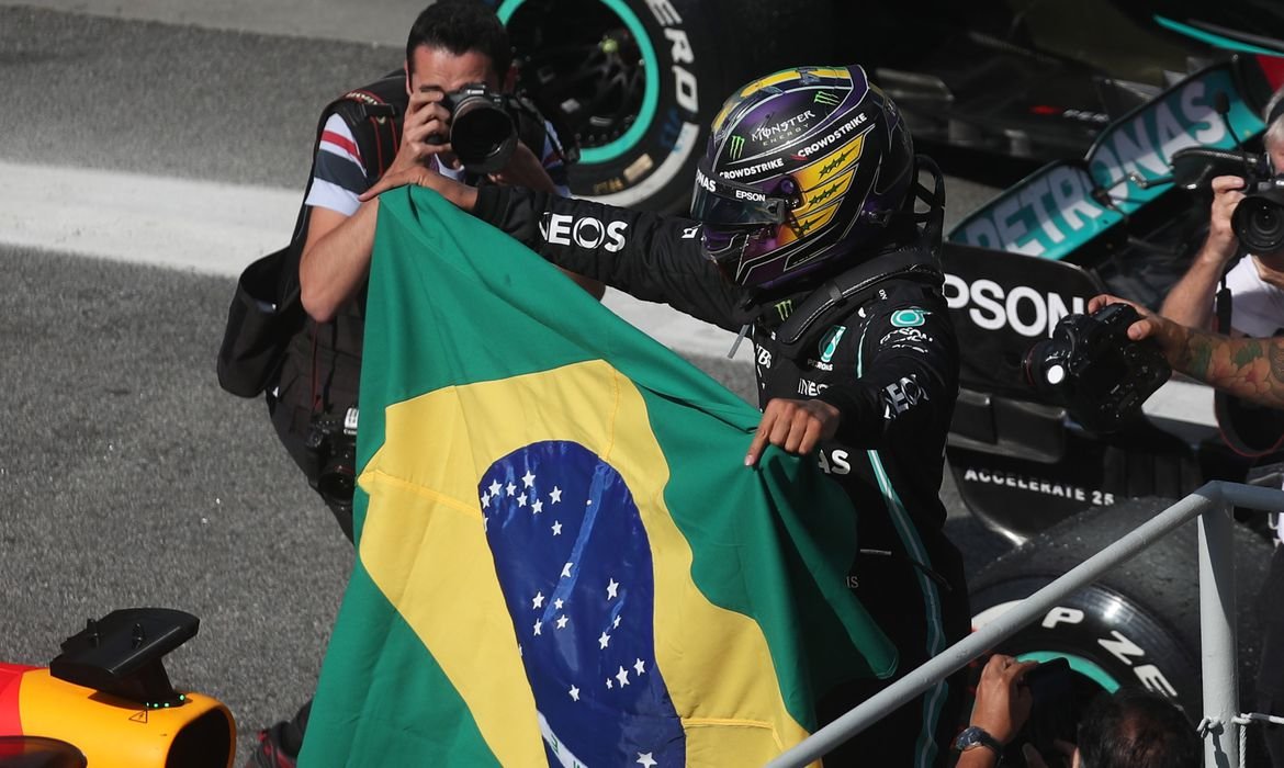 hamilton_vence_gp_de_sp #Pracegover foto: na imagem há duas pessoas, uma segurando a bandeira do Brasil e de capacete e a outra com uma máquina fotográfica