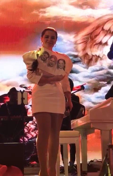 Maraísa #Pracegover foto: na imagem há uma mulher segurando um cartaz com a imagem de três pessoas