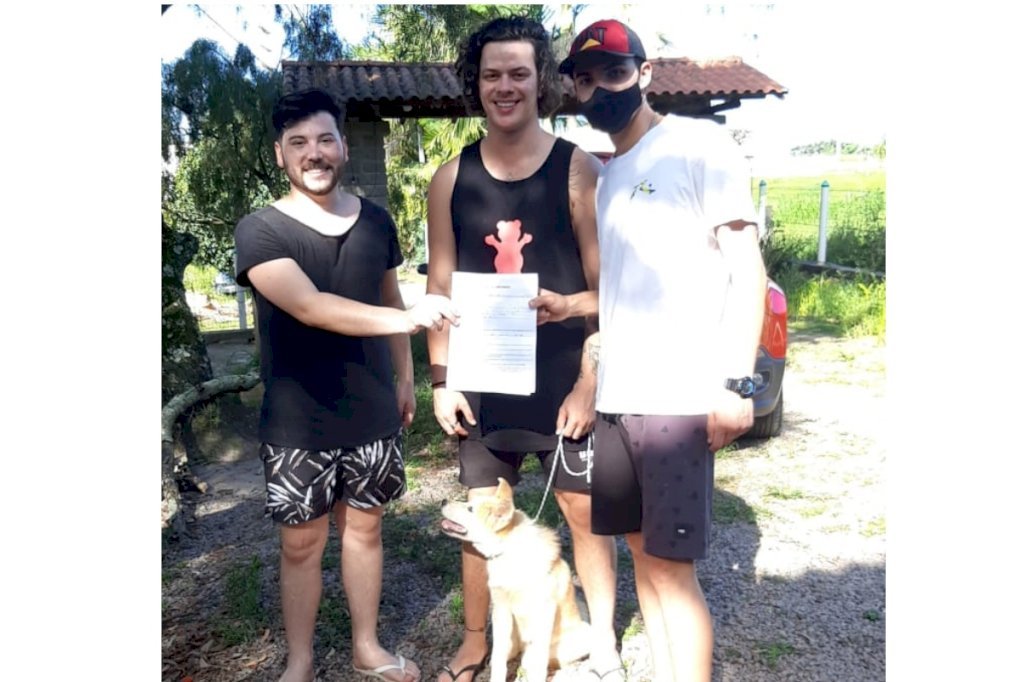 grupo #Pracegover Foto: na imagem há três jovens, um documento, um cão e árvores