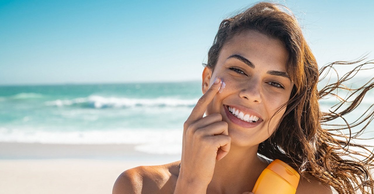 Mulher passando protetor solar em seu rosto (1) #Pracegover foto: na imagem há uma mulher, um frasco de protetor solar, o mar e areia