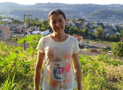 Roseli #Praceover foto: na imagem há uma mulher de camiseta clara, ela está sorrindo. Há casas, montanha e uma área verde