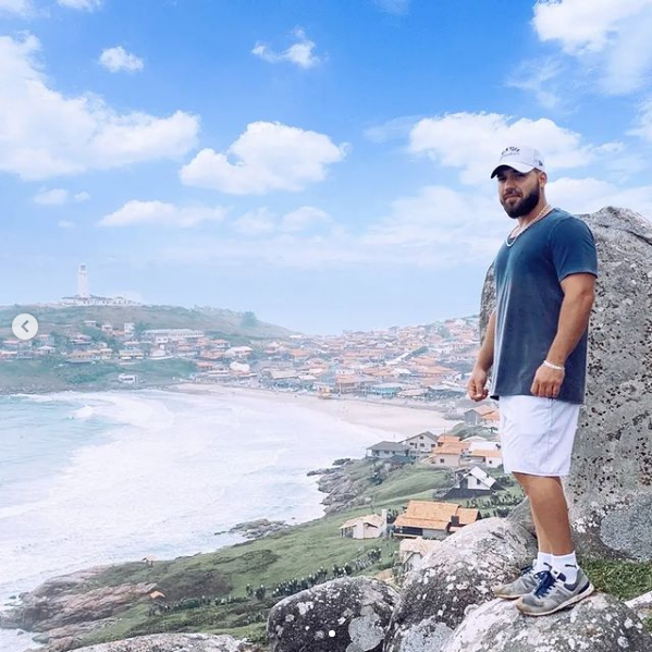 Captura de Tela 2022-01-14 às 23.12.36 #Pracegover Foto: na imagem há um homem de bermuda nranca, camiseta azul e boné claro. Há o mar, montanhas, pedras e casas