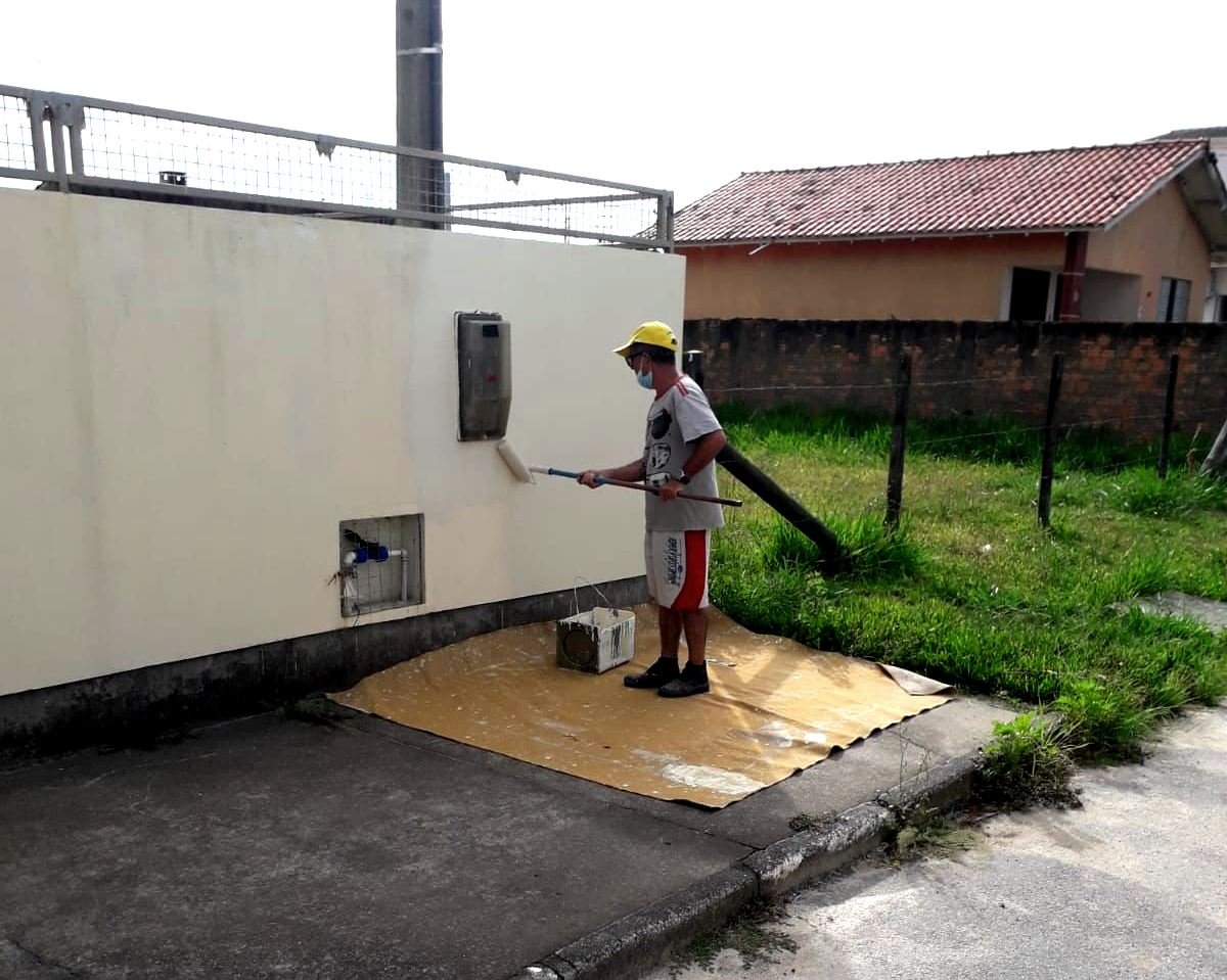 manutenção #Pracegover Foto: na imagem há um muro, calçada, um homem pintando o muro, residências e um terreno baldio