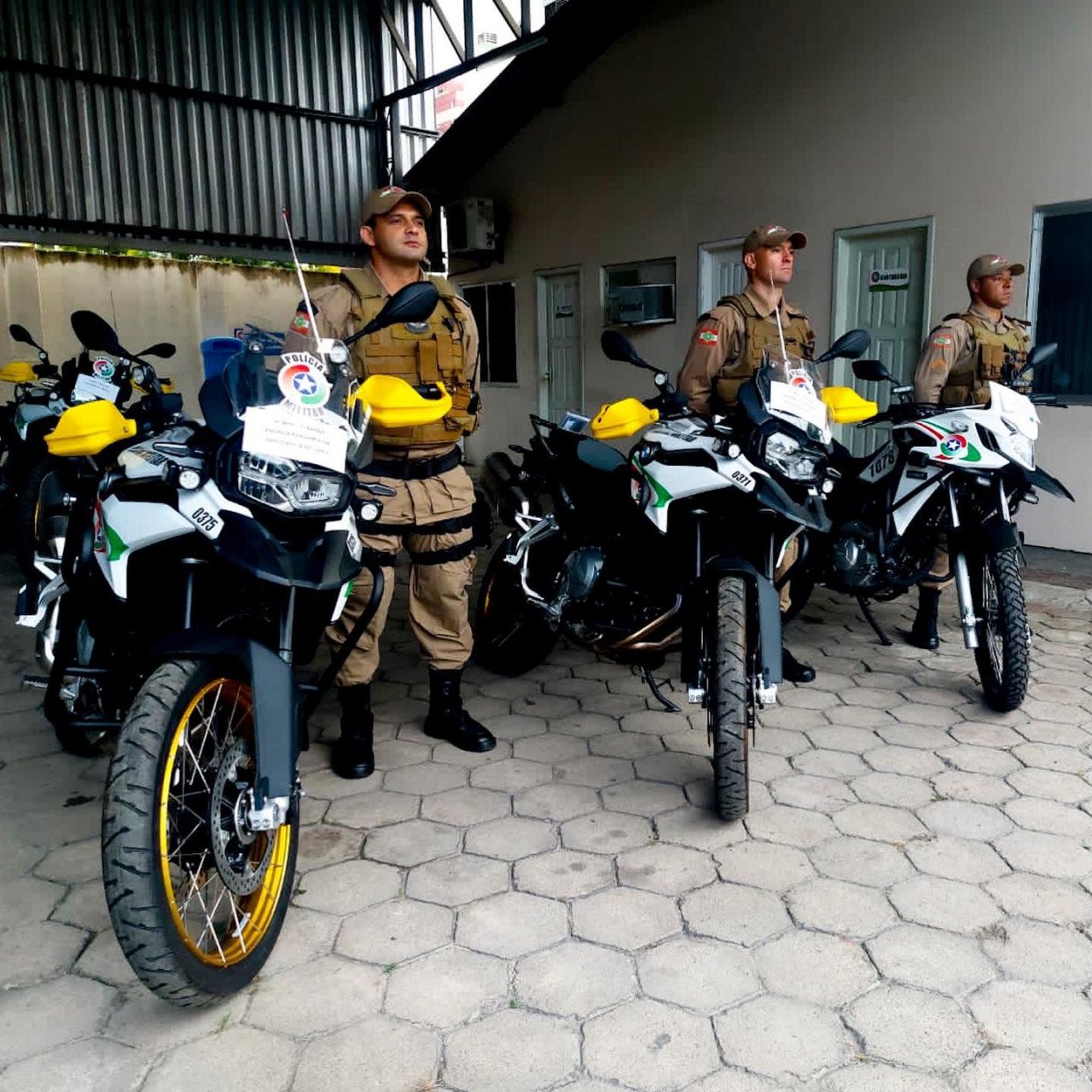 916bde46-55b3-4f38-bdf1-a44367148613 #PraCegoVer Na foto, três policiais militares estão parados ao lado de três motocicletas