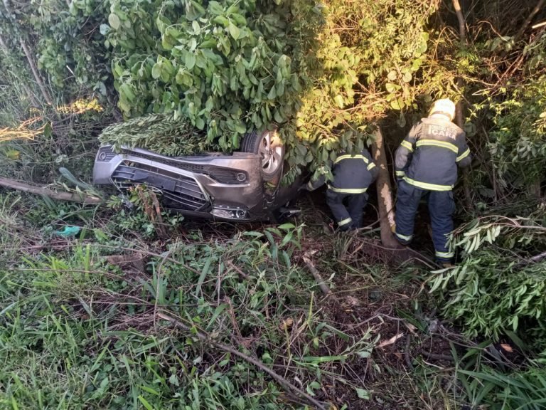 #PraCegoVer Na foto, há um carro capotado no meio do mato e dois bombeiros militares avaliam a situação