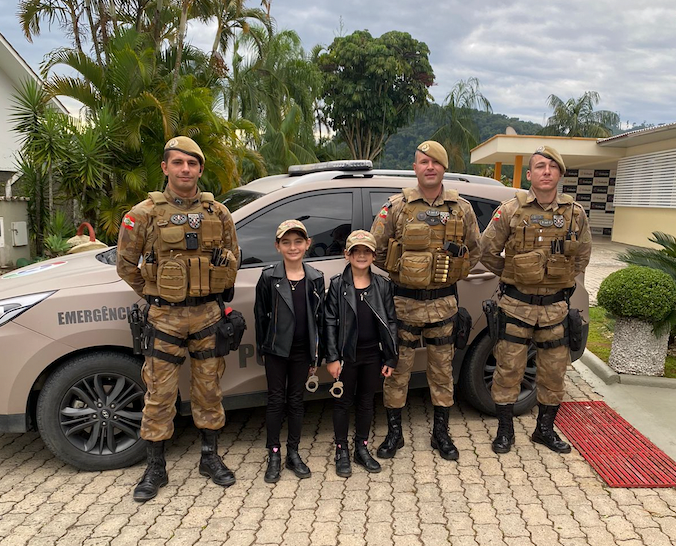 PM na escola #PraCegoVer Na foto, há uma viatura da Polícia Militar e, na frente, há três policiais e duas meninas