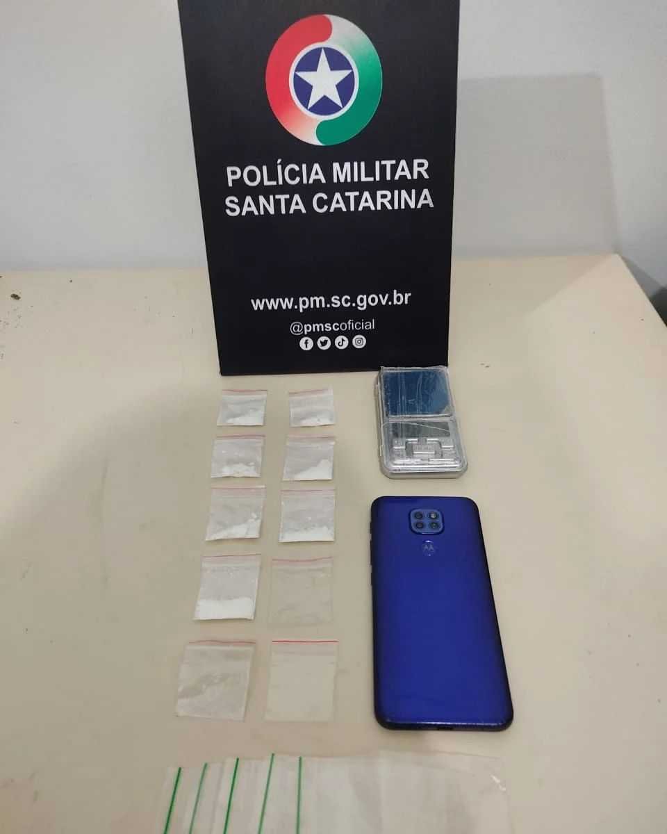 Droga Praia do Rosa #PraCegoVer Na foto, uma placa com o símbolo da Polícia Militar, drogas, uma balança de precisão e um aparelho de telefone celular