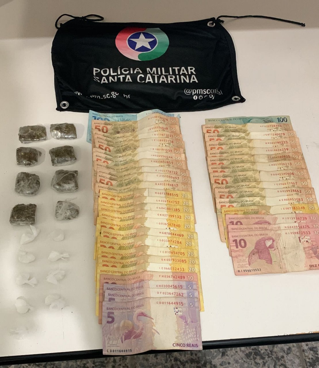 Droga Tubarão #PraCegoVer Na foto, uma bandeirola da Polícia Militar, diversas cédulas de dinheiro e porções de drogas
