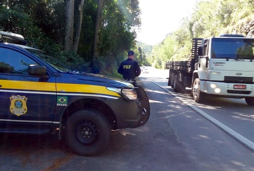 PRF 1 #PraCegoVer Na foto, um policial está ao lado de uma viatura e fiscaliza o trânsito em uma rodovia