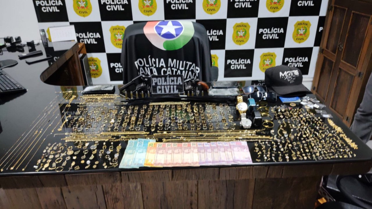 joalheria sombrio #PraCegoVer Na foto, uma mesa com vários produtos roubados. Ao fundo o símbolo da Polícia Civil e uma bandeira da Polícia Militar
