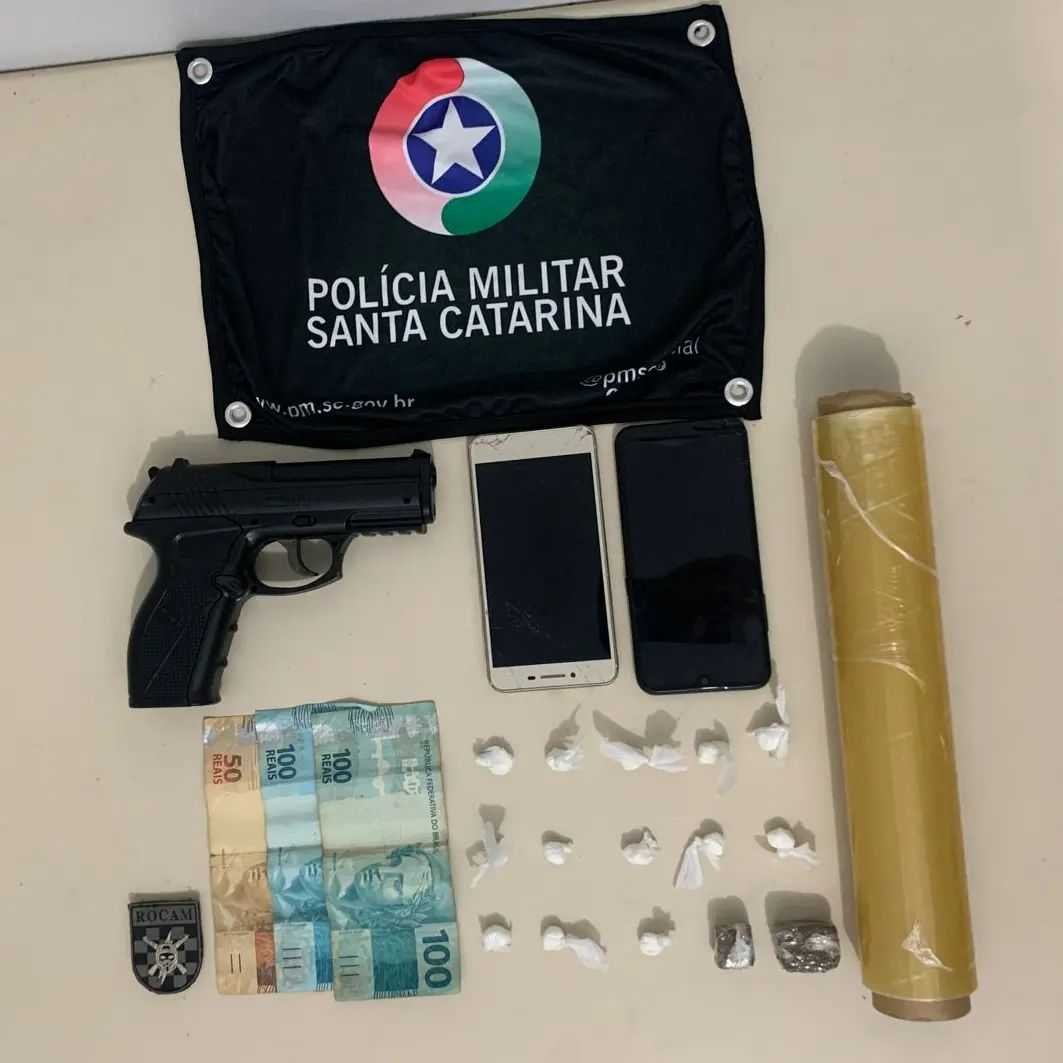 Arma Falsa - Imbituba #PraCegoVer Na foto, em cima de uma mesa há uma bandeirola com o símbolo da Polícia Militar, porções de drogas, cédulas de dinheiro, apetrechos usados para porcionar entorpecentes, dois aparelhos celulares e uma arma de fogo falsa