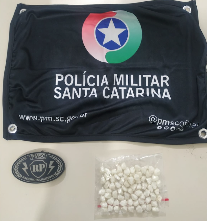 Crack em TB #ParaTodosVerem A foto mostra uma mesa e em cima dela há uma bandeirola com o símbolo da Polícia Militar, um pacote com pedras de crack e o brasão da Radiopatrulha da PM
