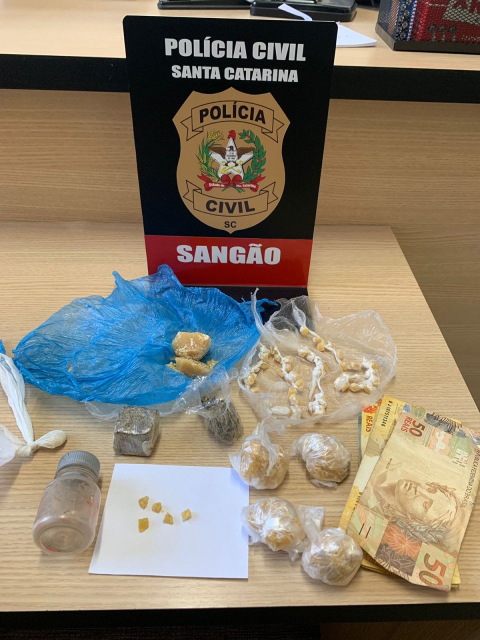 Droga Sangão #PraCegoVer Na foto, uma mesa onde há uma placa com o emblema da Polícia Civil de Sangão, cédulas de dinheiro e porções de crack e maconha