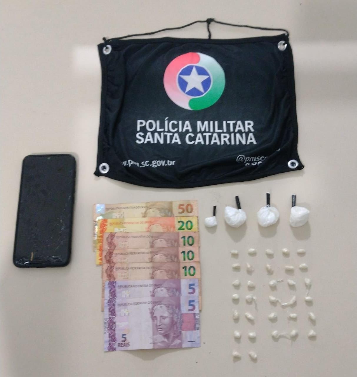 Droga TB #ParaTodosVerem Na foto, em cima de uma mesa hão símbolo da Polícia Militar de Santa Catarina, um aparelho de telefone celular preto, notas de dinheiro e porções de cocaína e crack