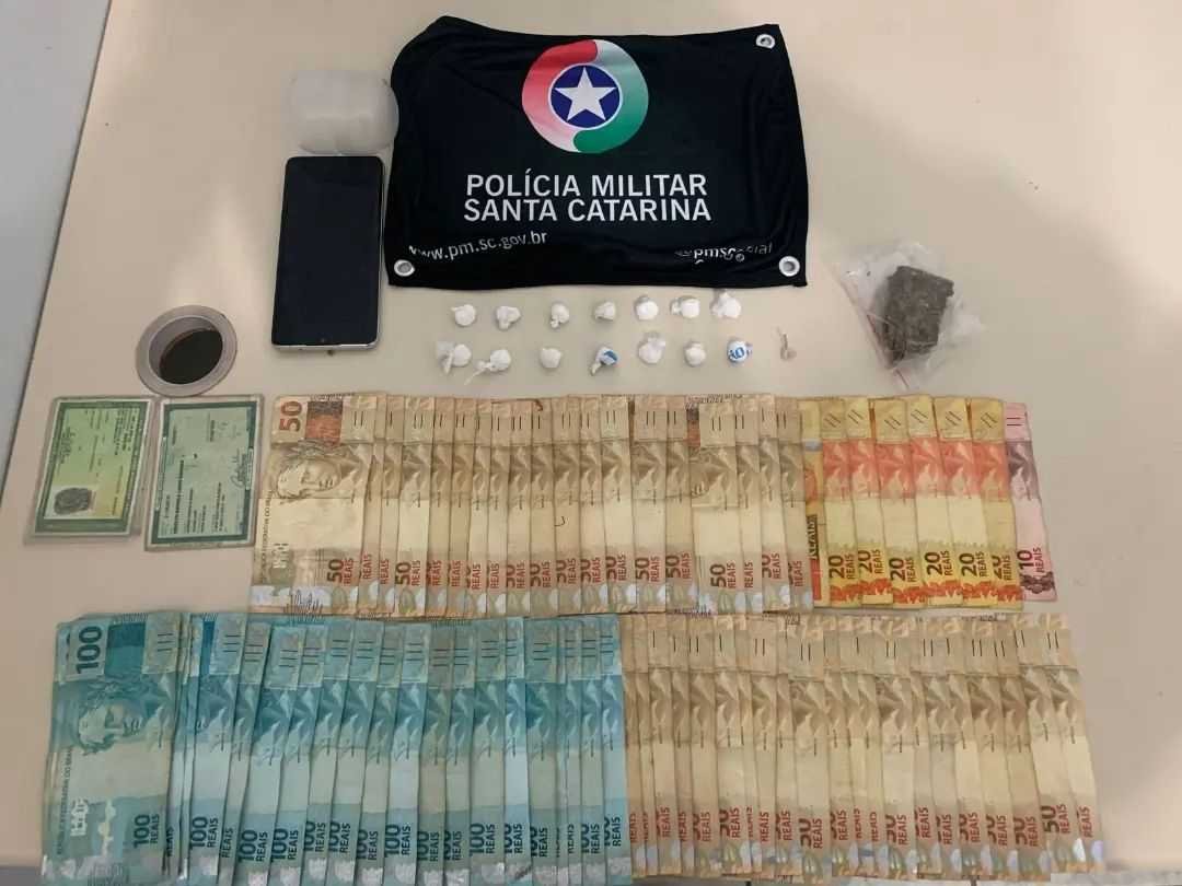Furto e tráfico Imbituba #ParaTodosVerem Na foto, em cima de uma mesa há o símbolo da Polícia Militar de Santa Catarina, porções de cocaína e maconha, um aparelho de telefone celular preto, diversas cédulas de dinheiro e dois documentos de identificação